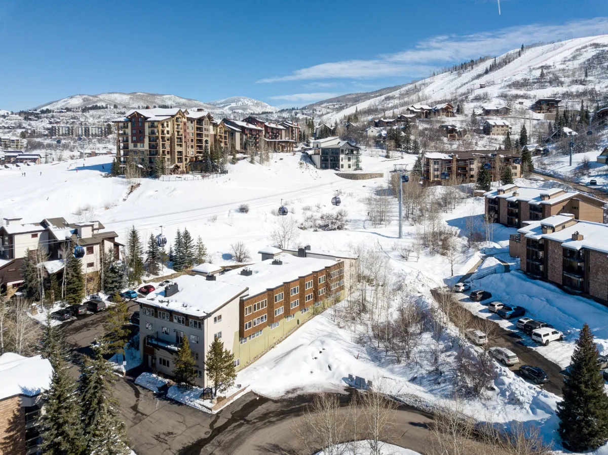 A serene morning image featuring snow-covered hills, modern buildings, pine trees, and a parking area. The backdrop of mountains and open sky sets the tone for peaceful yet active living, with hints of year-round activities like skiing, hiking, and biking.