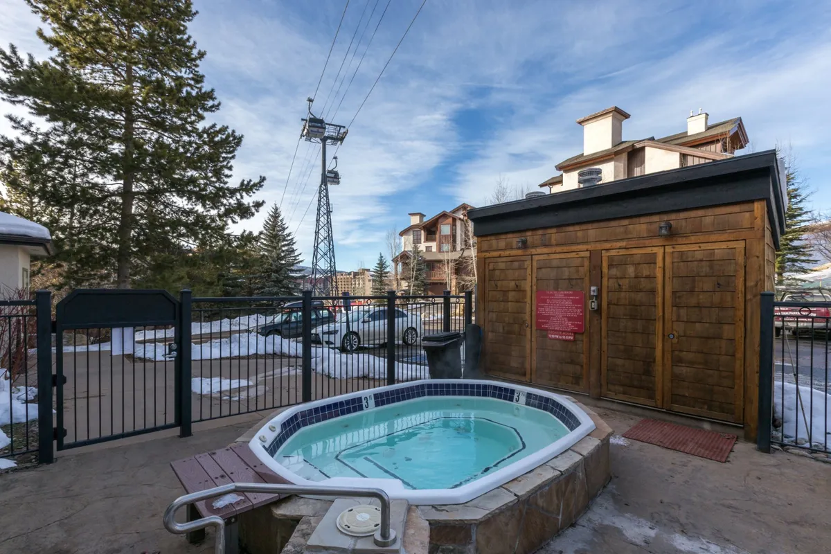 An inviting outdoor hot tub sits next to a quaint wooden shed, all surrounded by a snowy landscape. The bubbling water contrasts beautifully with the cool winter air, and a ski lift is visible in the background, emphasizing the resort setting and mountain access.