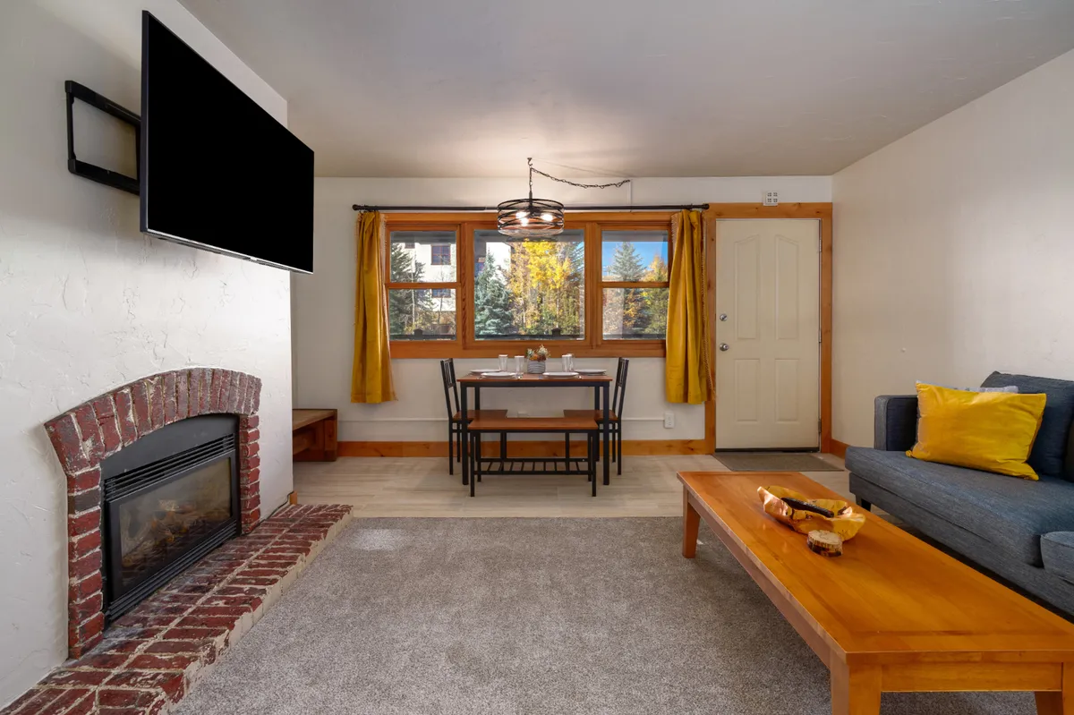 This interior scene features a dining table near a large window, with a view toward a cozy fireplace and an adjacent sofa. The setting is warm and welcoming, ideal for shared meals or relaxed evenings.
