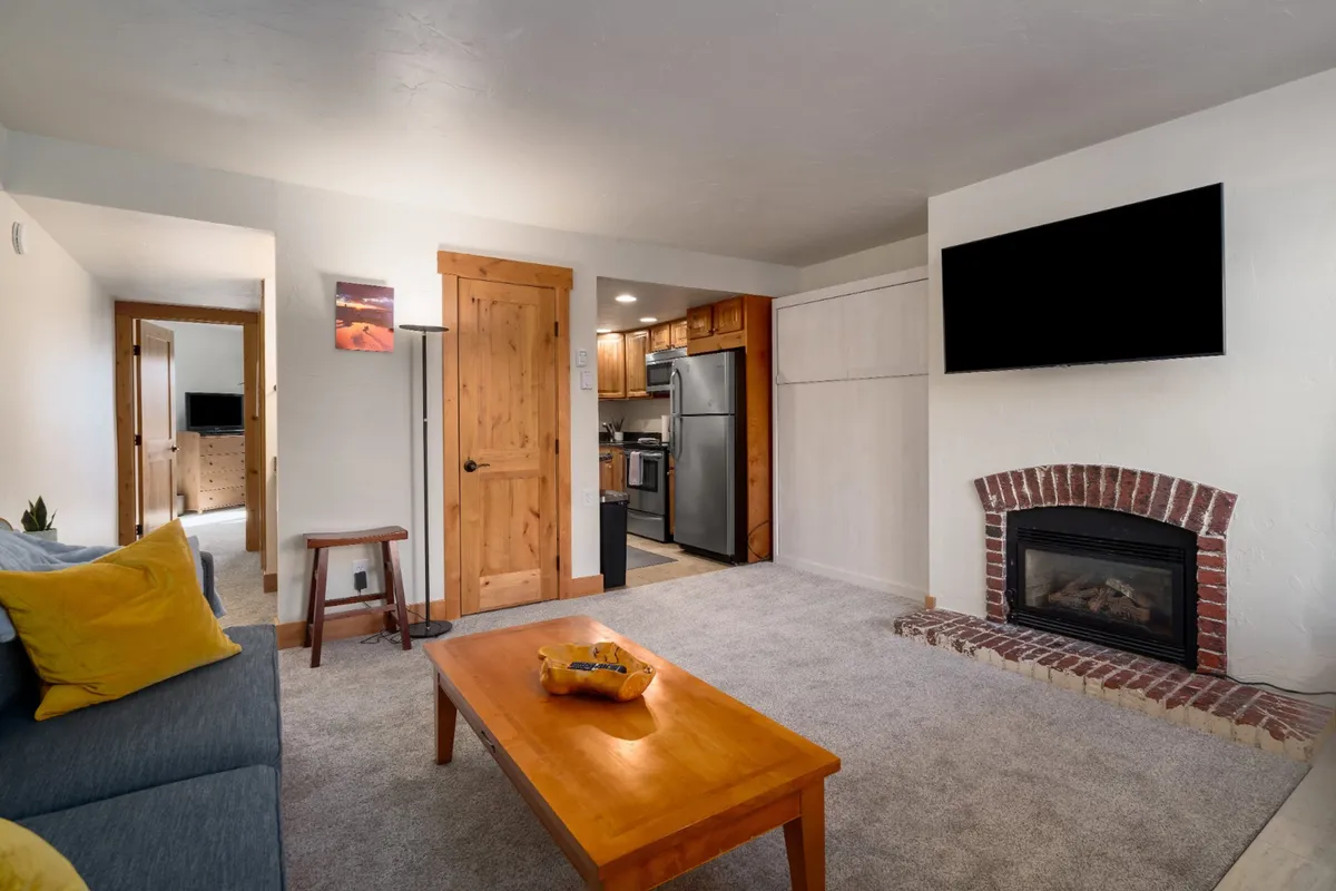 A comfortable interior scene with a plush sofa adorned with a bright yellow pillow, a rustic brick fireplace, a wooden coffee table, and a kitchen area visible in the background. The setup emphasizes spaces for both relaxing and socializing, with clear visual connections between comfort and gathering.