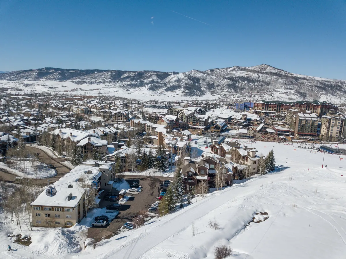 A wide view of the Steamboat ski resort area blanketed in fresh snow, with the bustling town set against the dramatic backdrop of snow-covered mountains. The scene captures lively winter sports activity, parked cars indicating easy access, and various buildings hosting local restaurants and shops.