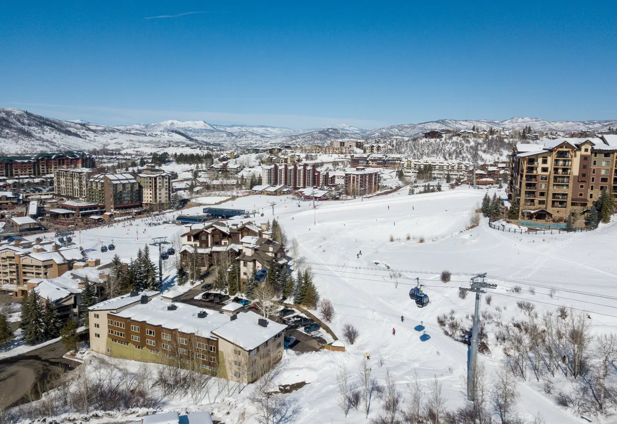 A scenic panorama of snow-blanketed mountains, open ski slopes, charming chalets, a gondola lift, and the heart of the town center. The golden light of sunset adds warmth, while visible shops and restaurants hint at après-ski gatherings and local exploration.