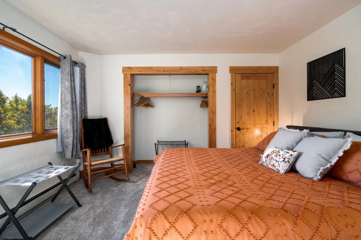 This image showcases a bedroom with a bed covered in orange bedding, a large window that fills the space with natural light, a classic wooden rocking chair, and a closet with hangers. The inviting setup highlights the room's warmth and tranquil ambiance.