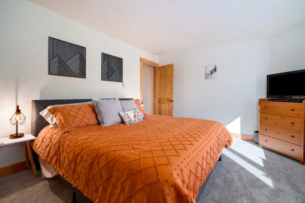 A warm and inviting bedroom scene featuring a bed with an orange quilt, decorative pillows, wall art, a nightstand with a lamp, and a sturdy wooden dresser. The arrangement exudes a feeling of comfort, perfect for restful nights or leisurely mornings.