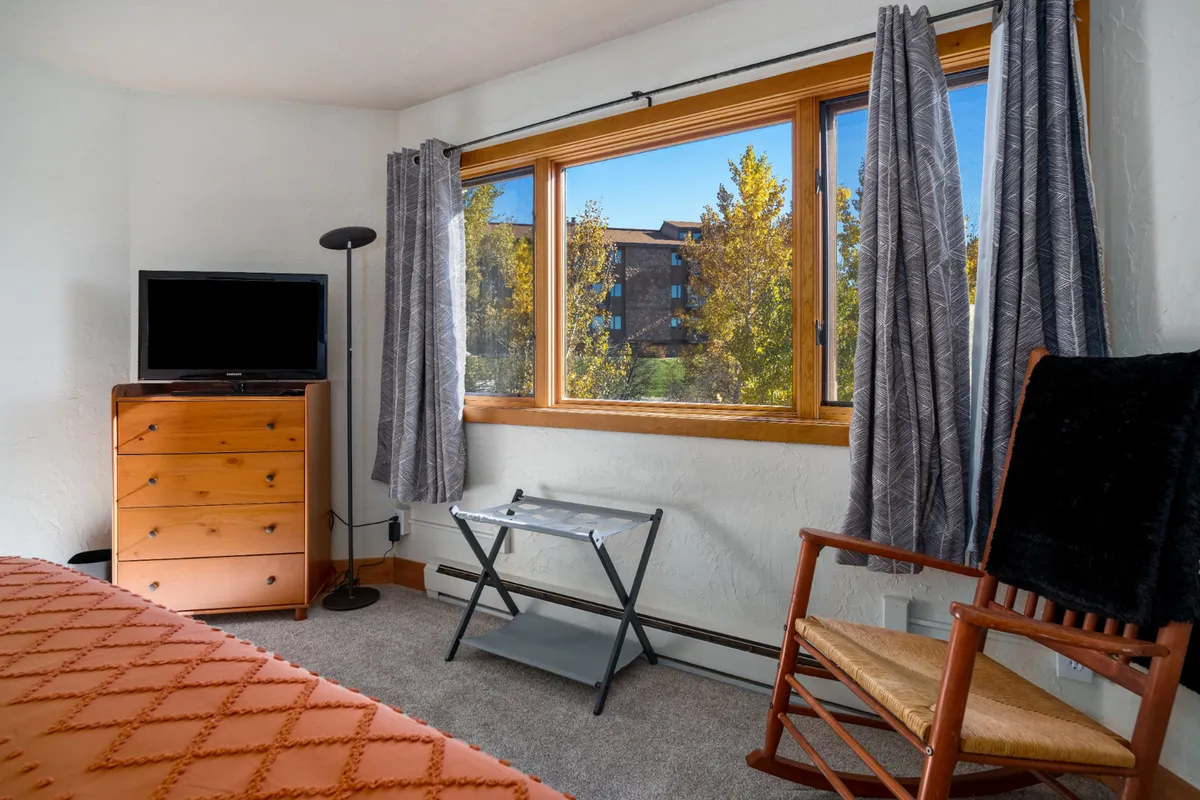 The image focuses on a spacious bedroom area with a large window offering views outside, a wooden dresser for storage, a television set for entertainment, and a comfortable rocking chair placed near the window. The overall scene is serene and inviting.