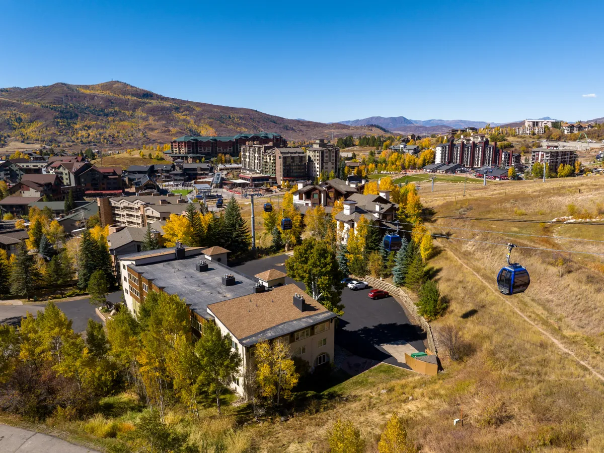A panoramic view of resort buildings nestled at the base of majestic mountains, with a gondola lift rising into the sky and colorful trees in the foreground. The outdoor scene highlights the accessibility of both winter and summer mountain activities.