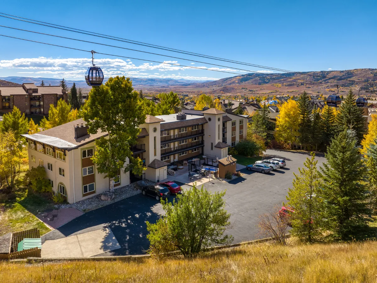 The image showcases the exterior of a charming condo building nestled among towering trees, with a nearby gondola lift hinting at immediate ski access. Snow-capped mountains loom in the distance, and a well-maintained parking area offers convenience. The scene radiates a sense of adventure and mountain serenity, perfect for outdoor enthusiasts.