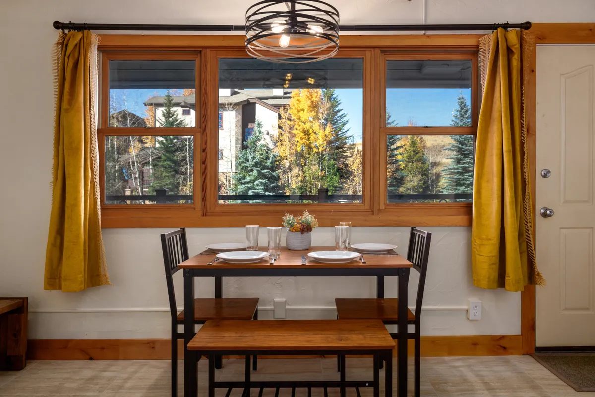 A cozy dining area with a wooden dining table occupies the foreground, surrounded by large windows framed with yellow curtains. A decorative centerpiece adds charm, and ample natural light streams in, creating an inviting atmosphere for meals.