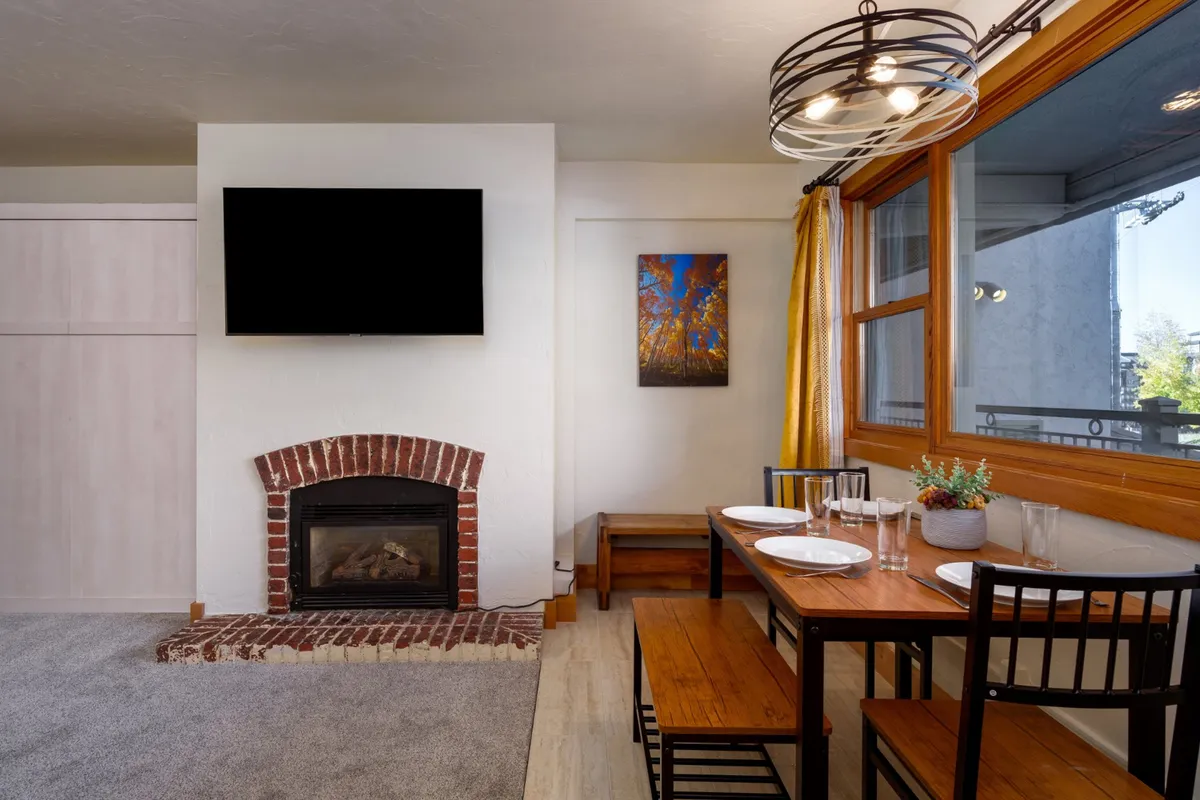 A warmly lit living space featuring a prominent gas fireplace as the focal point, surrounded by comfortable seating including a plush sofa. Large windows with curtains frame inviting views and natural light, while a dining table nearby adds to the social, communal feel of the room.